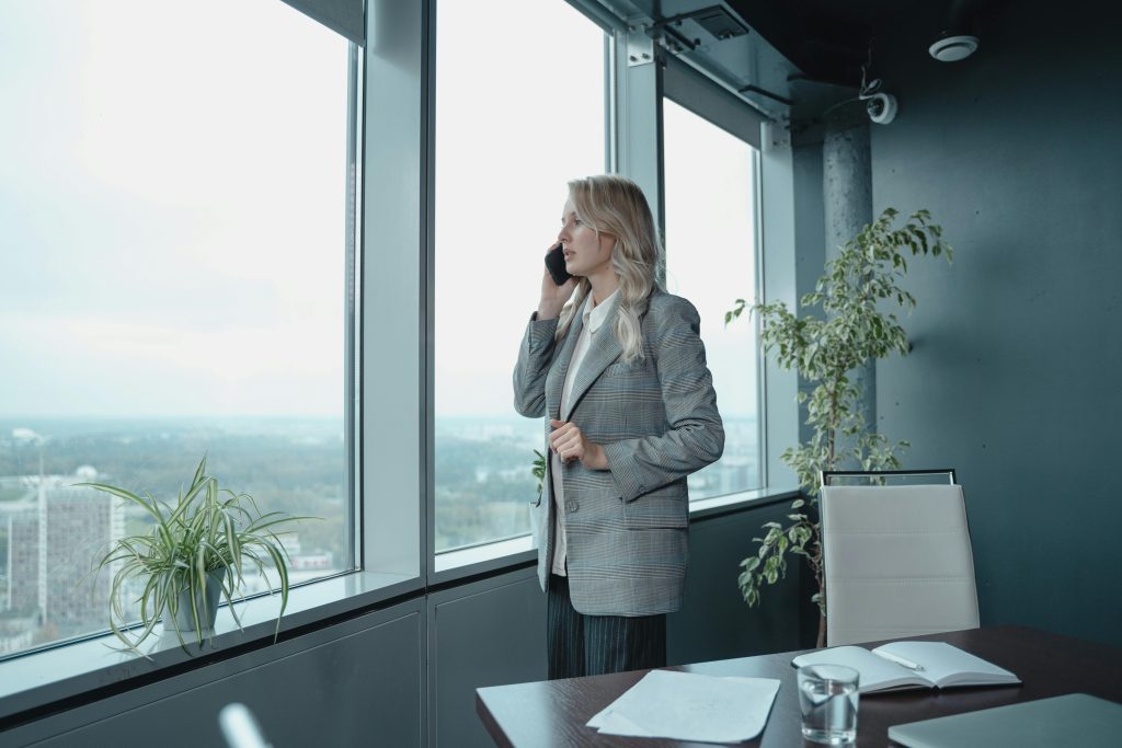 A professional woman in a gray blazer talks on the phone in a modern office with large windows.
